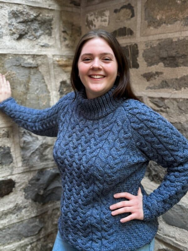 Smiling woman in an Irish Aran crewneck sweater with classic cable and diamond stitches, ivory wool, symbolizing luck and heritage.