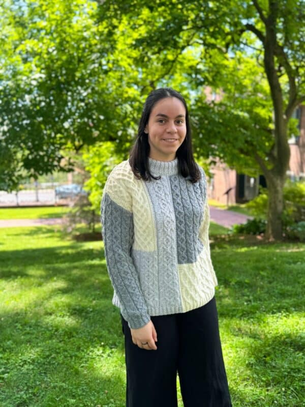 Irish woman with dark hair smiles on green grass, wearing a cream Aran crewneck sweater with classic cable knit patterns.