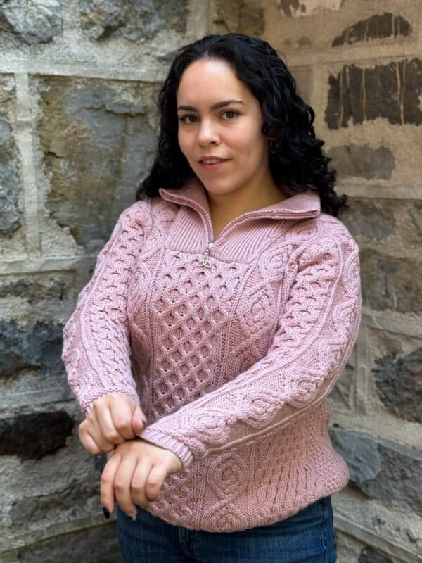 Irish woman in a pink wool Aran half zip sweater with classic cable knit by stone wall; heritage craftsmanship and warmth.