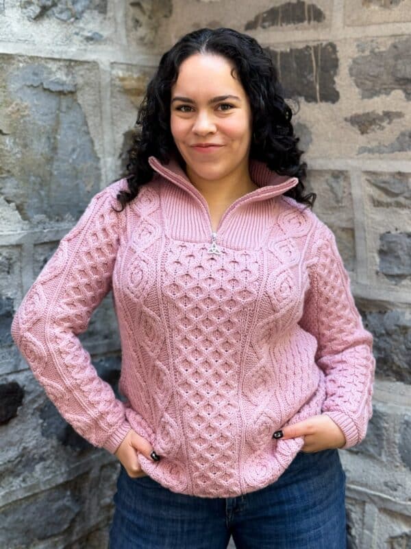 Irish woman with curly hair wears an Aran half zip sweater in cream wool, featuring classic cable knit by a stone wall.