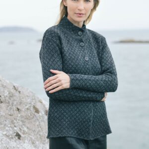 A woman in a Scottish tweed jacquard jacket and tartan skirt stands by the sea, arms crossed, against a rugged shore backdrop.