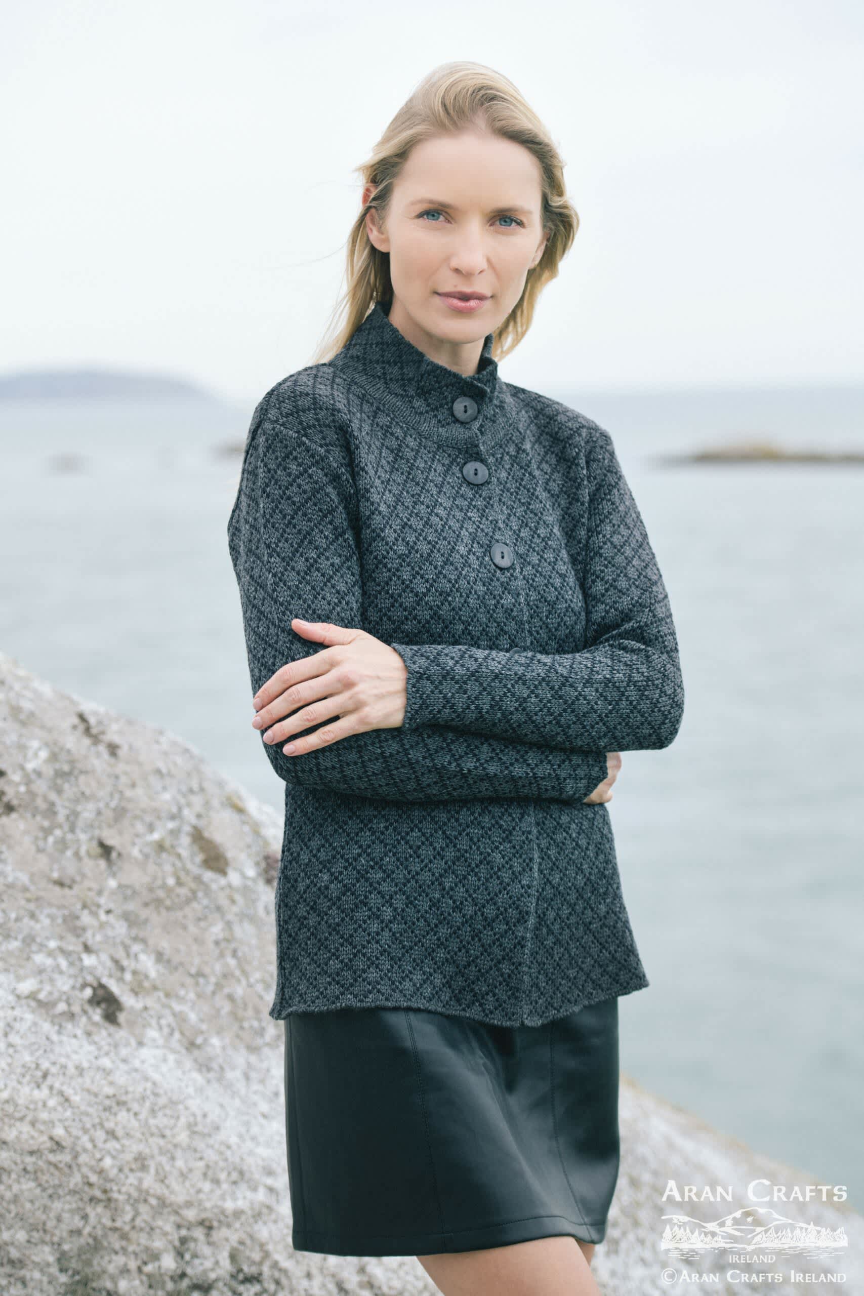 A woman in a Scottish tweed jacquard jacket and tartan skirt stands by the sea, arms crossed, against a rugged shore backdrop.