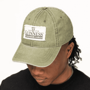 Irish-inspired olive green wool flat cap (Guin GN9039) with classic patchwork, worn by curly-haired person in black shirt.